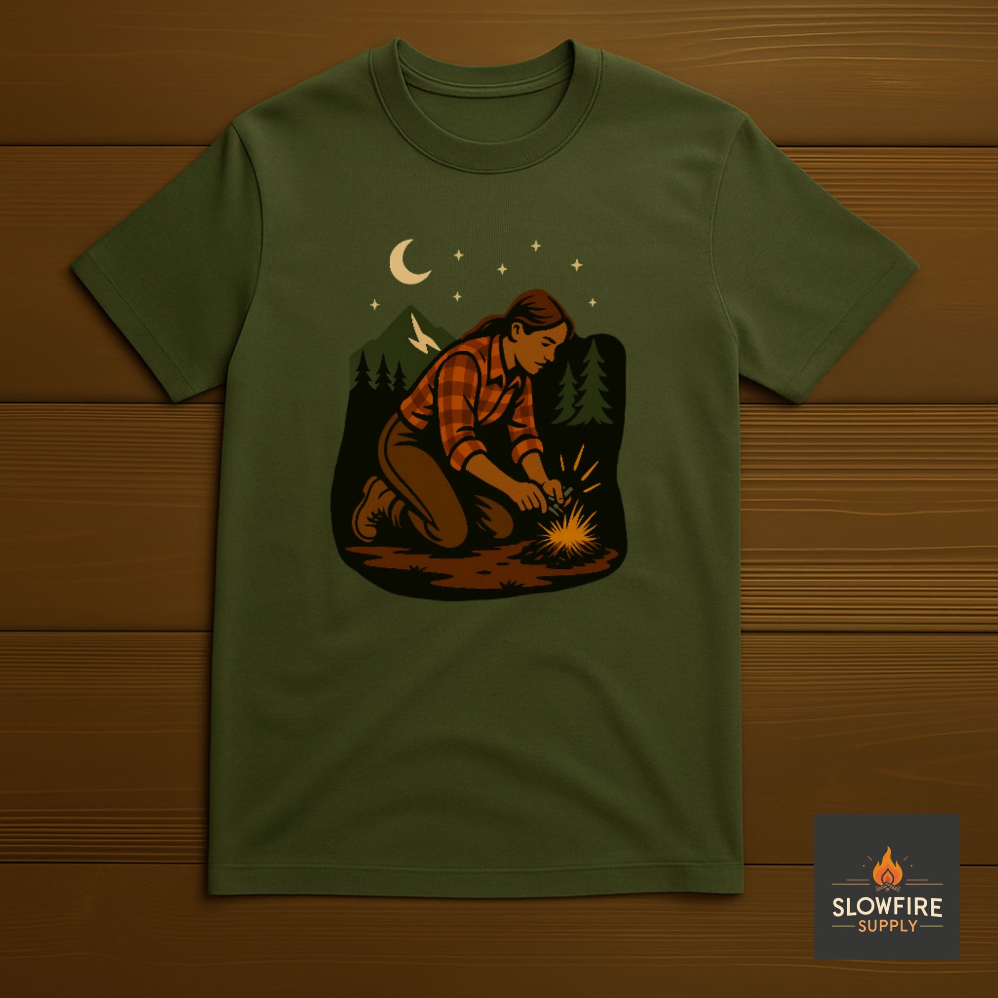 Green t-shirt with a graphic of a person building a fire in the woods, displayed on a wooden surface.