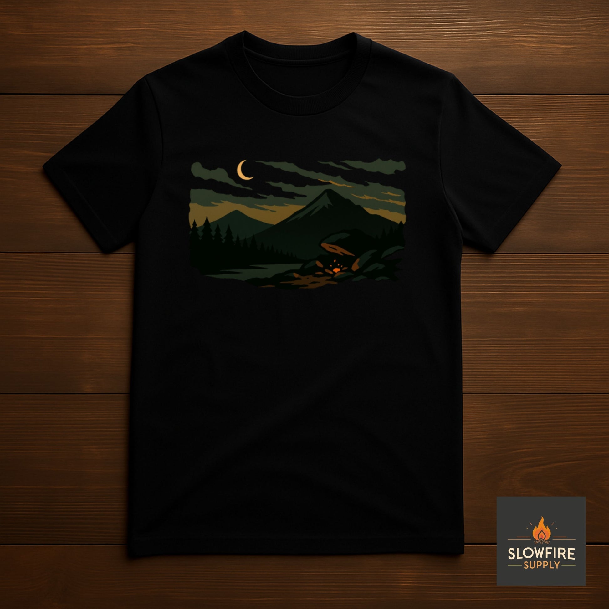 Black t-shirt with a mountain and campfire design on a wooden surface, featuring the Slowfire Supply logo.
