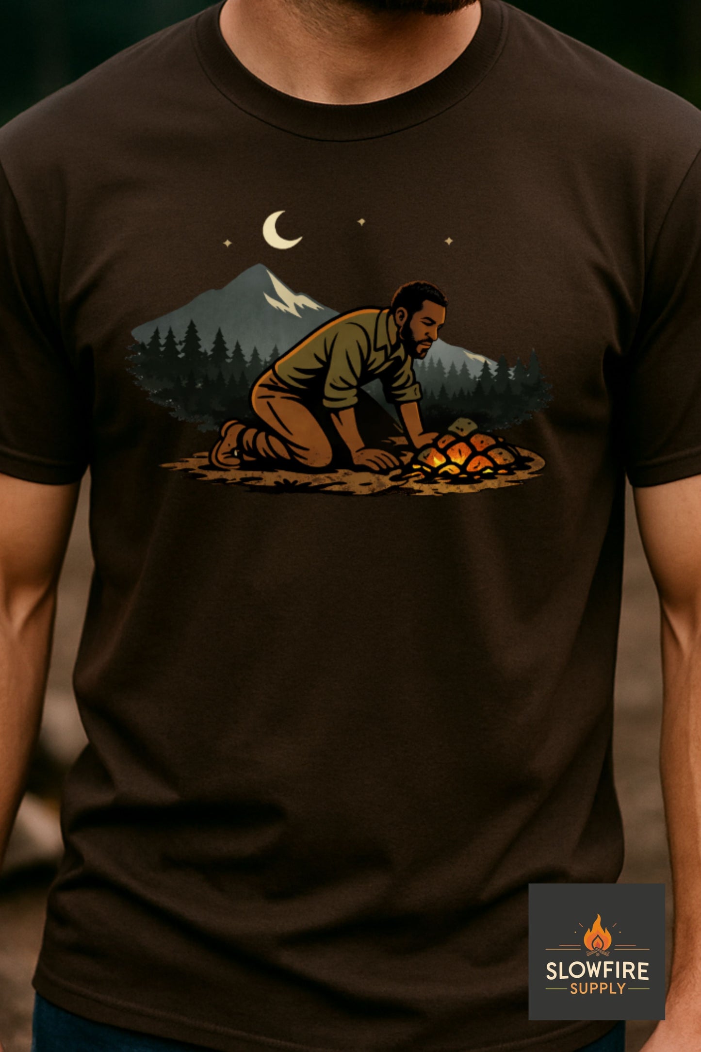 Brown t-shirt with a graphic of a person cooking over a campfire at night, worn by a person.