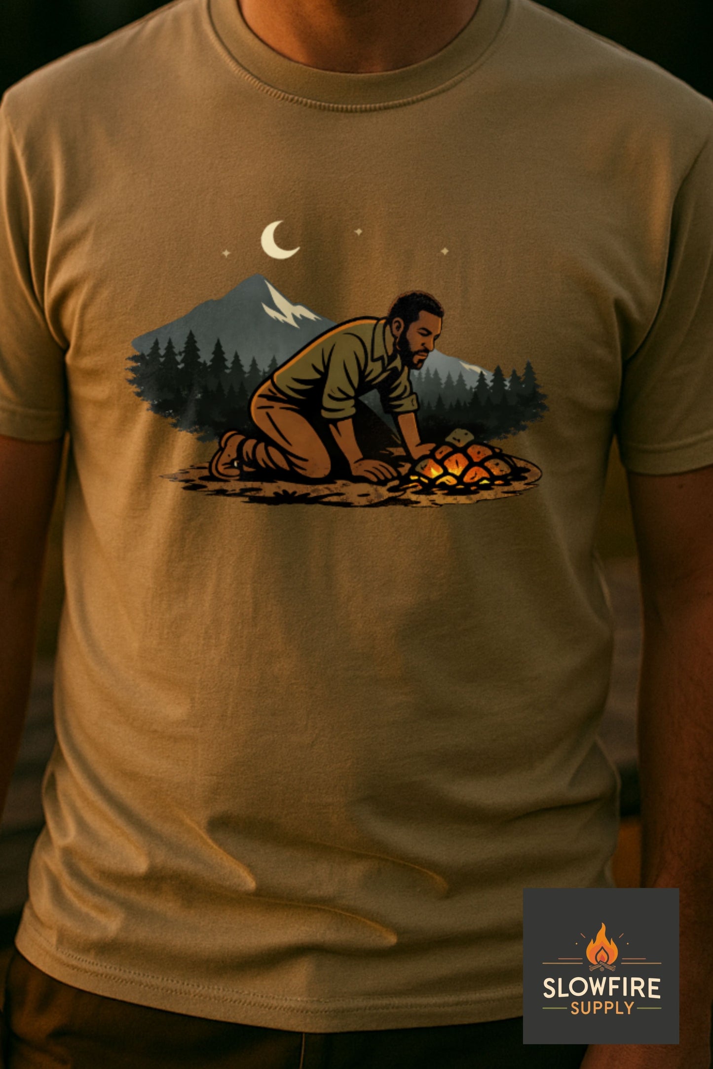 Beige t-shirt with a graphic of a person by a campfire and mountains, worn by a person.